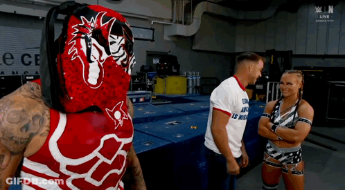 A Wrestler In A Red And White Outfit Is Talking To A Man And A Woman In A Room . GIF