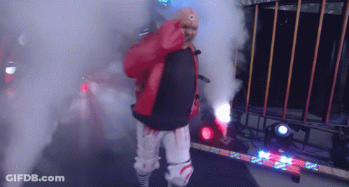 A Wrestler In A Red Jacket And White Pants Is Walking Through Smoke GIF