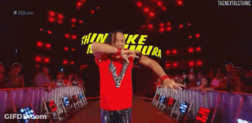 A Wrestler In A Red Shirt Is Kneeling Down In Front Of A Large Sign That Says Shinsuke Nakamura . GIF