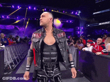 A Wrestler In A Studded Vest Stands In Front Of A Crowd At A Wrestling Match GIF