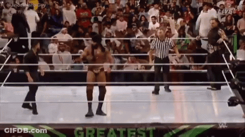 A Wrestler Is Dancing In A Wrestling Ring With A Referee In Front Of A Crowd . GIF