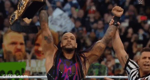 A Wrestler Is Holding A Championship Belt In The Air While A Referee Looks On . GIF