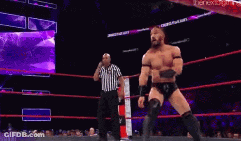 A Wrestler Is Jumping In The Air While Another Wrestler Is Standing Next To Him In A Wrestling Ring . GIF
