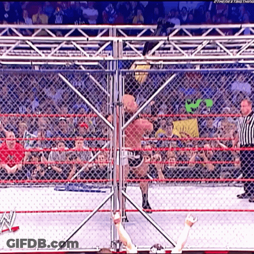 A Wrestler Is Jumping Over A Chain Link Fence While A Referee Looks On GIF