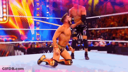 A Wrestler Is Kneeling Down In A Wrestling Ring While Another Wrestler Is Standing Behind Him . GIF