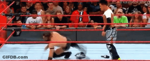 A Wrestler Is Kneeling Down In A Wrestling Ring While Another Wrestler Looks On . GIF