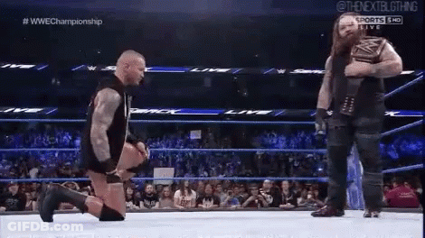 A Wrestler Is Kneeling Down In A Wrestling Ring While Another Wrestler Stands Behind Him . GIF