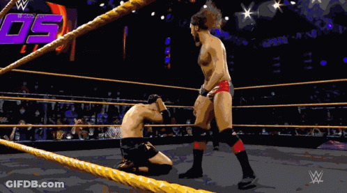 A Wrestler Is Kneeling Down In A Wrestling Ring While Another Wrestler Stands In The Corner GIF