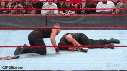 A Wrestler Is Kneeling Down Next To Another Wrestler Who Is Laying On The Floor . GIF
