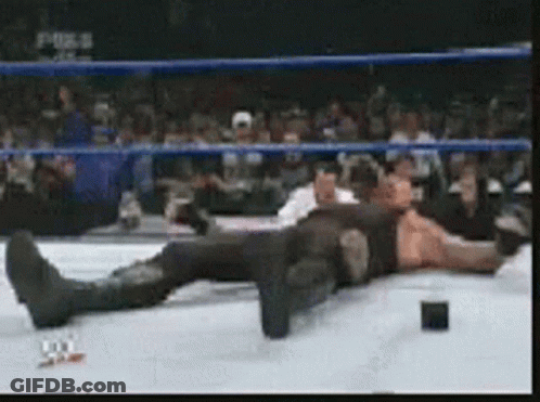 A Wrestler Is Laying On The Ground In A Wrestling Ring While A Crowd Watches GIF