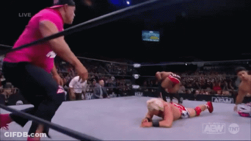 A Wrestler Is Laying On The Ground In A Wrestling Ring While A Man In A Pink Shirt Looks On . GIF