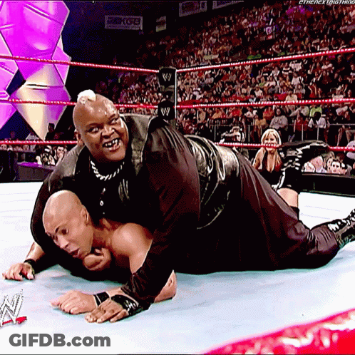 A Wrestler Is Laying On The Ground While Another Wrestler Holds Onto Him GIF