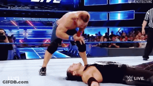 A Wrestler Is Laying On The Ground While Another Wrestler Is Standing On Top Of Him . GIF