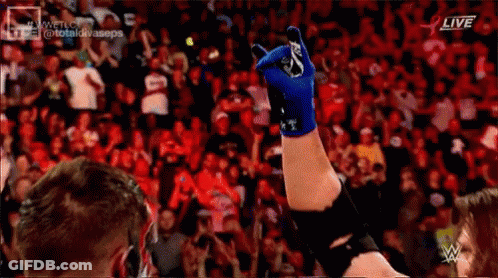A Wrestler Is Raising His Hand In The Air In Front Of A Crowd While Wearing A Blue Glove . GIF