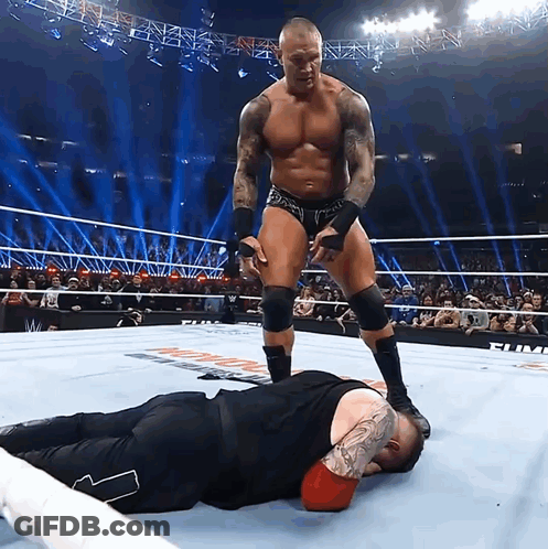 A Wrestler Standing Over Another Wrestler Laying On The Floor GIF