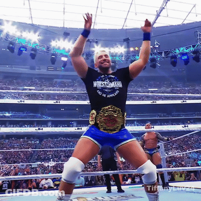 A Wrestler Wearing A Wrestlemania Shirt Stands In A Ring With His Arms In The Air GIF