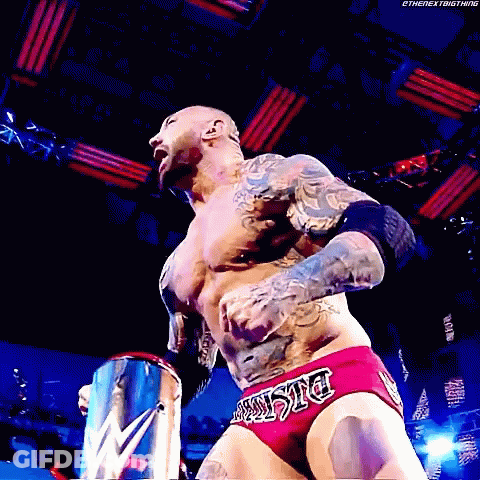 A Wrestler With A Tattoo On His Chest Is Wearing A Pair Of Red Trunks With The Word Justice On The Side GIF