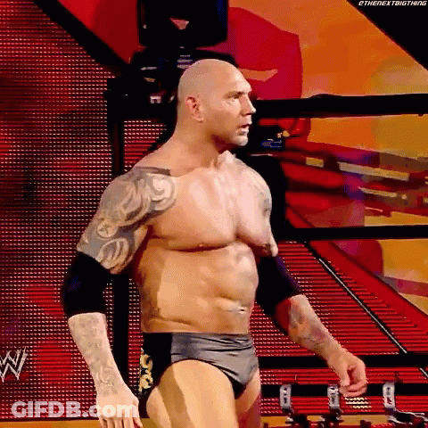 A Wrestler Without A Shirt Is Standing In A Ring With A Red Background . GIF