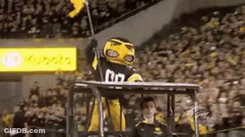 A Yellow And Black Mascot Is Sitting On Top Of A Tractor Holding A Flag . GIF