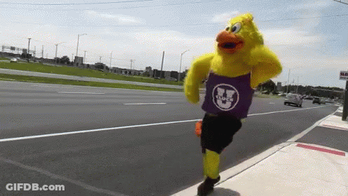 A Yellow Duck Mascot Wearing A Purple Shirt With A Smiley Face On It Is Running Down A Street GIF