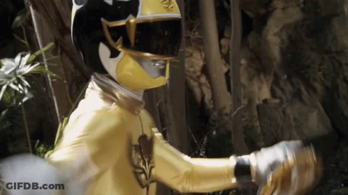 A Yellow Ranger With A Black Helmet And Gloves Is Standing In The Woods . GIF