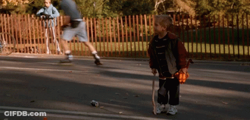 A Young Boy Holding A Cane Stands On The Side Of A Road GIF