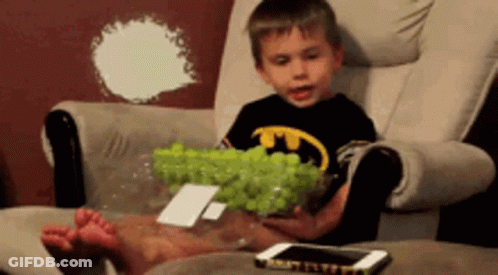 A Young Boy In A Batman Shirt Is Sitting In A Chair Holding A Bowl Of Green Grapes GIF
