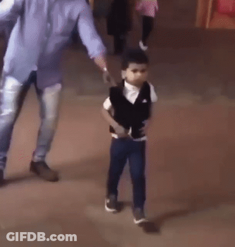 A Young Boy In A Black Vest And White Shirt Is Standing On A Sidewalk . GIF