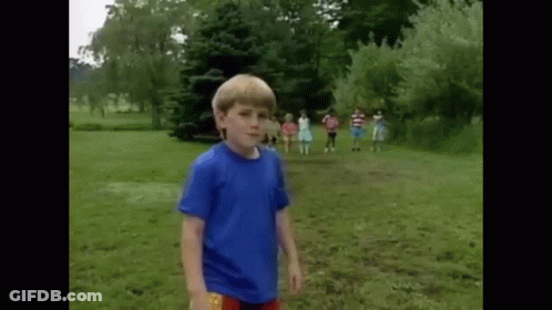 A Young Boy In A Blue Shirt Is Standing In A Grassy Field With A Group Of Children In The Background . GIF
