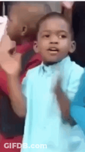 A Young Boy In A Blue Shirt Is Standing In Front Of A Group Of People . GIF