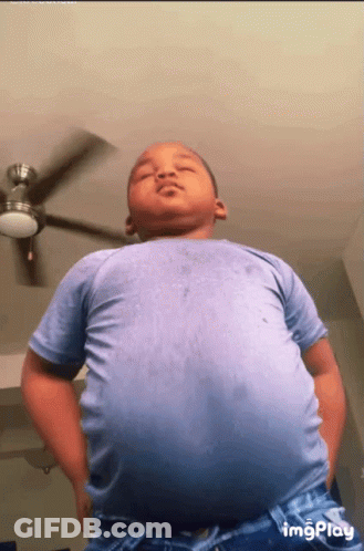 A Young Boy In A Blue Shirt Is Standing Under A Ceiling Fan GIF