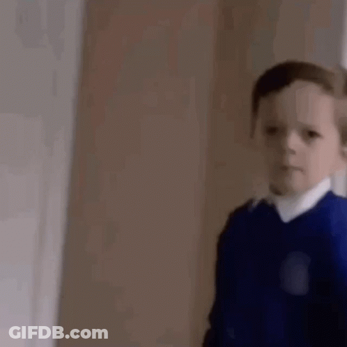 A Young Boy In A Blue Sweater And White Shirt Is Standing In A Room . GIF