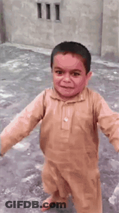 A Young Boy In A Brown Shirt Is Smiling And Walking On A Sidewalk . GIF