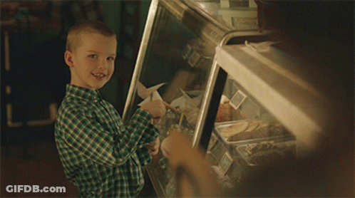 A Young Boy In A Green Plaid Shirt Is Smiling In Front Of A Display Case Of Food GIF