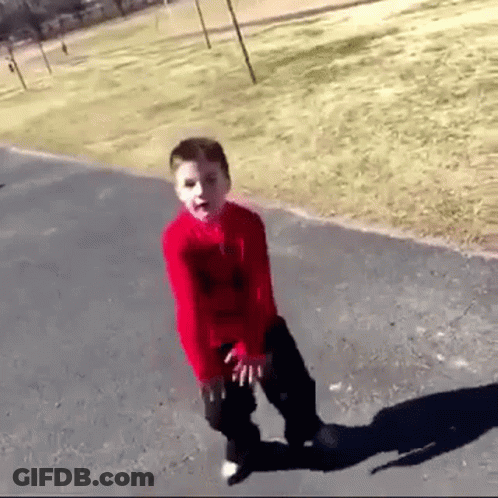 A Young Boy In A Red Shirt And Black Pants Is Standing On A Sidewalk . GIF