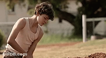 A Young Boy In A Tank Top Is Digging In The Dirt In A Field . GIF