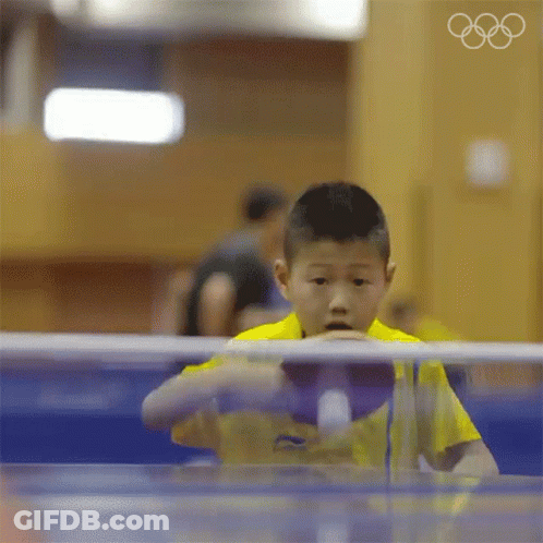 A Young Boy In A Yellow Shirt Is Playing Ping Pong With The Olympic Rings In The Background GIF