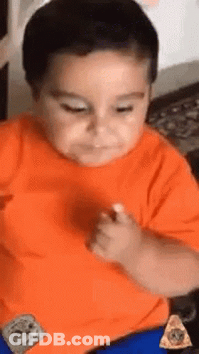 A Young Boy In An Orange Shirt Is Sitting At A Table With A Slice Of Pizza . GIF