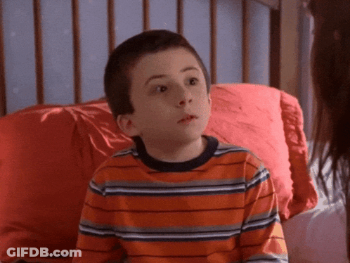 A Young Boy In An Orange Striped Shirt Sits On A Bed With A Red Pillow GIF