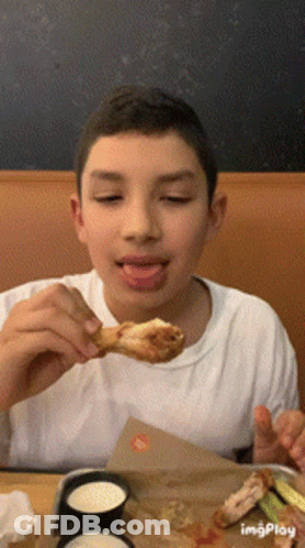 A Young Boy Is Eating A Chicken Wing With His Tongue Hanging Out GIF
