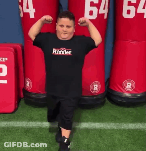 A Young Boy Is Flexing His Muscles On A Football Field In Front Of A Row Of Red Dummies . GIF