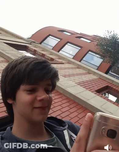 A Young Boy Is Holding A Cell Phone In Front Of A Building GIF
