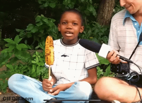 A Young Boy Is Holding A Corn On The Cob And Talking Into A Microphone GIF