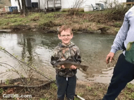 A Young Boy Is Holding A Fish In His Hands In Front Of A Stream . GIF