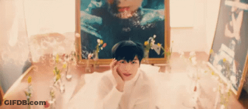 A Young Boy Is Laying In A Bathtub With Flowers And A Picture Of A Woman In The Background . GIF