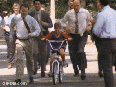 A Young Boy Is Riding A Bike With A Group Of Men Following Behind Him GIF