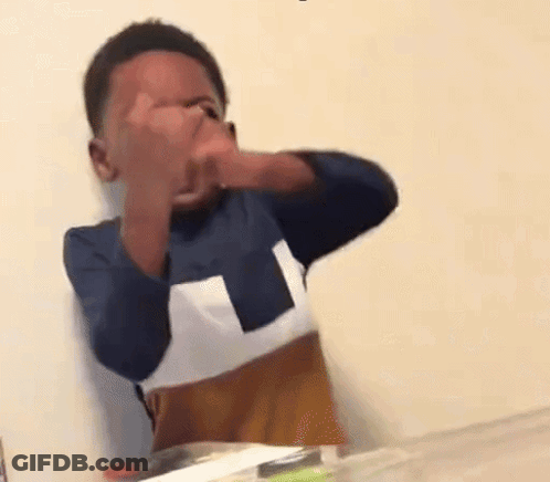 A Young Boy Is Sitting At A Table Covering His Face With His Hands . GIF