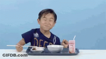 A Young Boy Is Sitting At A Table Eating Food With Chopsticks And A Strawberry Milk . GIF