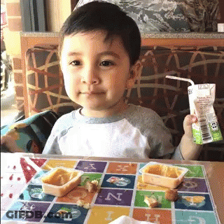 A Young Boy Is Sitting At A Table Holding A Carton Of Juice With A Straw GIF