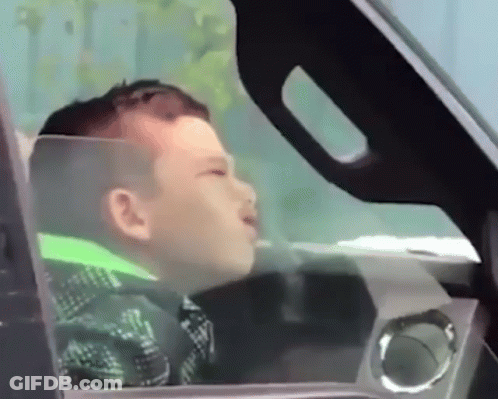 A Young Boy Is Sitting In The Driver 'S Seat Of A Car Looking Out The Window GIF